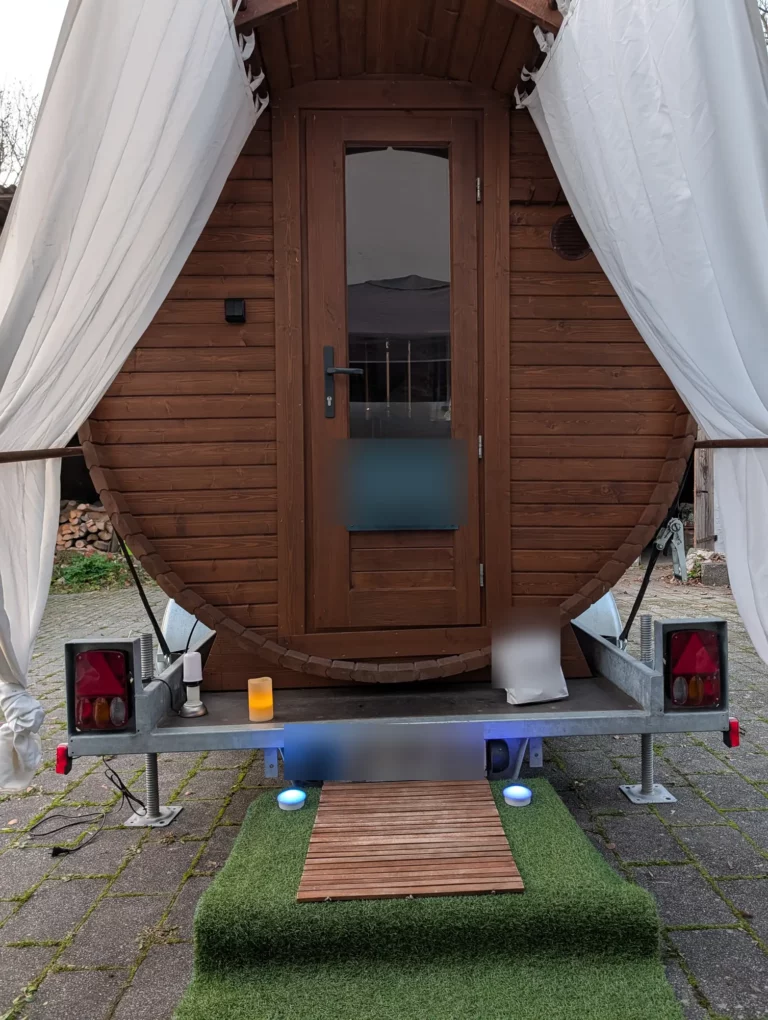 Eingang der Fasssauna mit Vorhängen, Holzsteg und Kunstrasen
